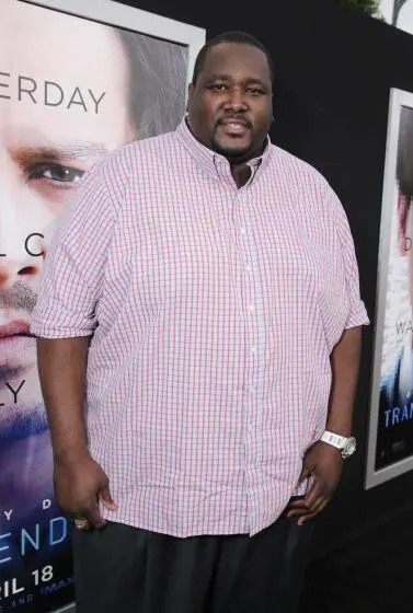 Quinton Aaron in a red and white patterned button down shirt posing at a red carpet.