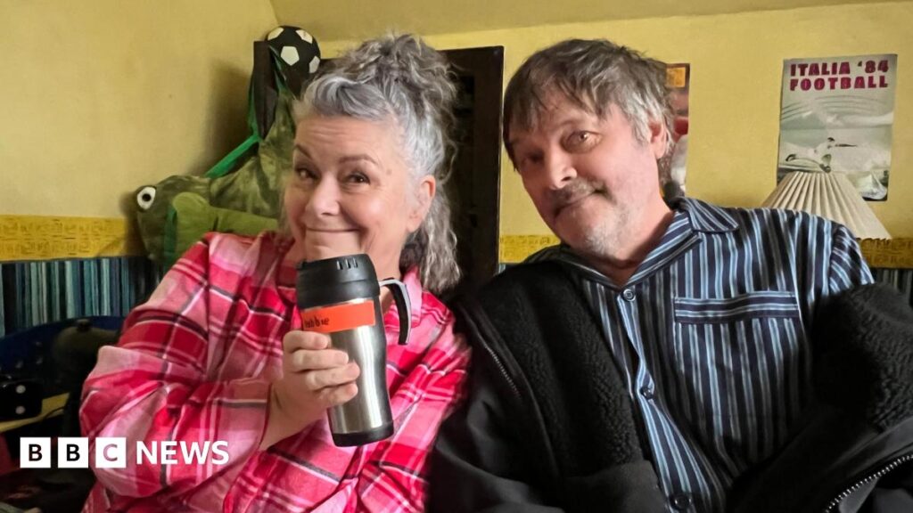 Dawn French sat next to Mark Heap on a sofa. They are smiling at the camera.