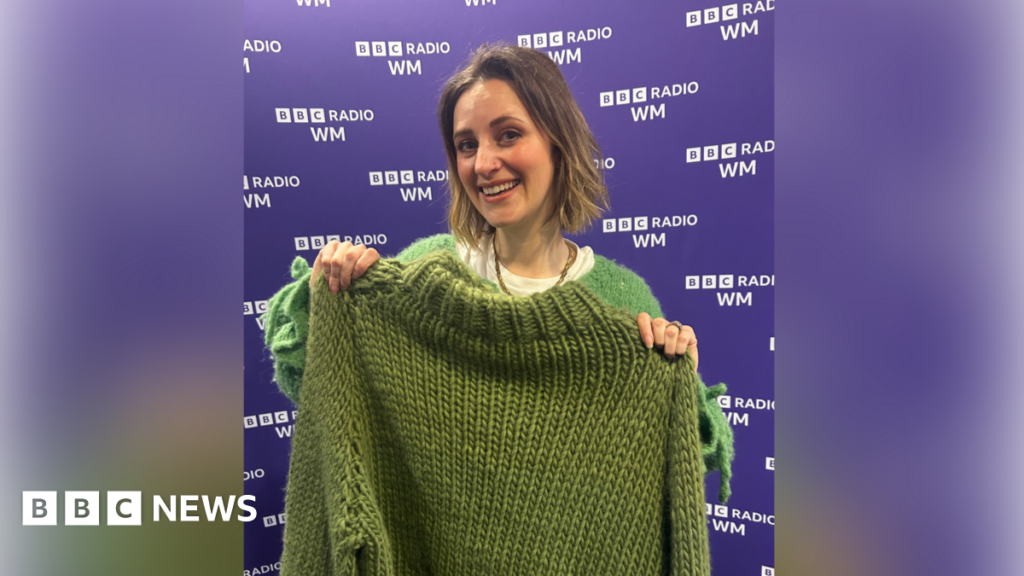 A woman with short brown hair grins at the camera as she holds up a green knitted jumper. She stands in front of a purple BBC Radio WM background.