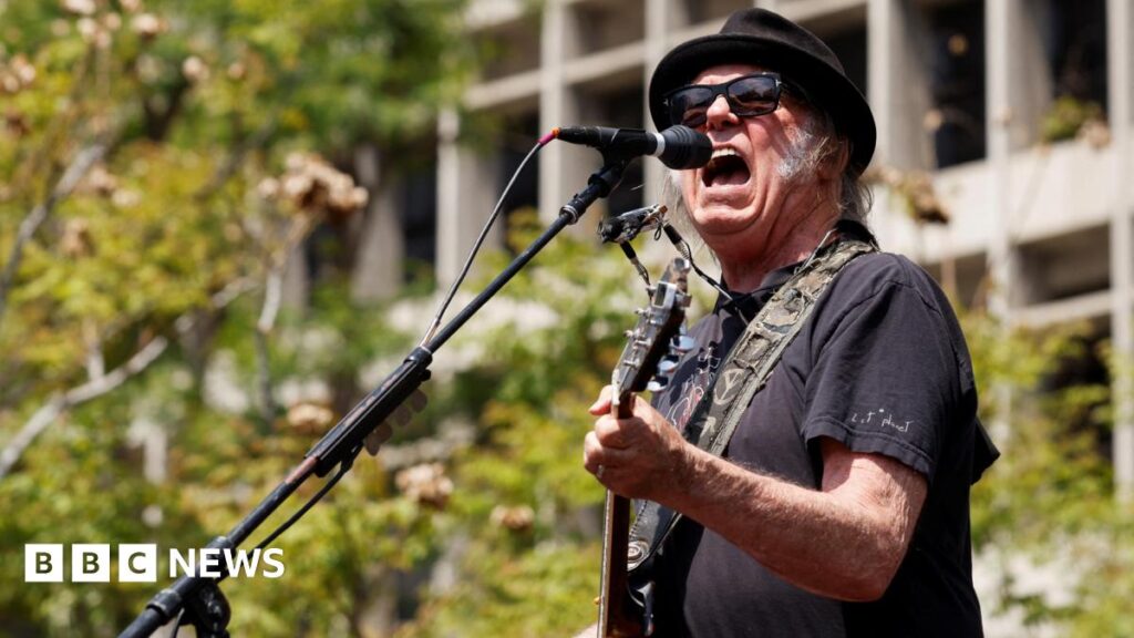 The rock star Neil Young plays a guitar and sings on a stage outside in the daytime.