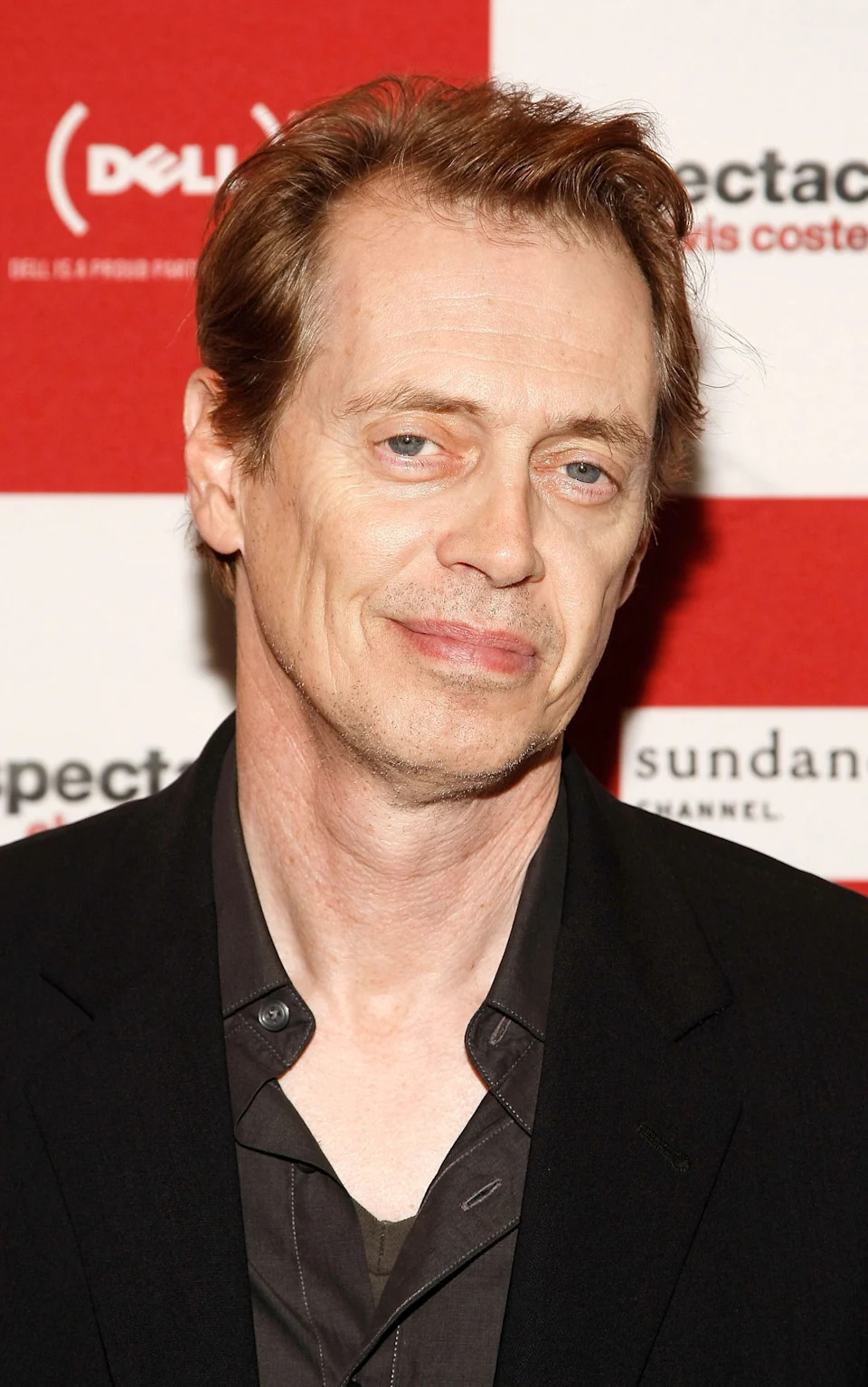 Person smiling on the red carpet, wearing a black suit with a partially unbuttoned shirt