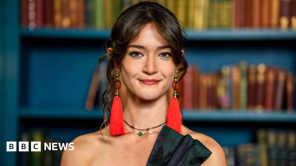 'Outpouring of support' for Warwick Traitors student Jade Scott – BBC A head and shoulders photo of a young woman with dark hair tied back, with loose curls around her face. She is wearing red earrings with long tassels, a green jewelled choker necklace and the top of a tartan bow on a shoulderless corset can just be seen. Behind her are blue painted shelves with books.
