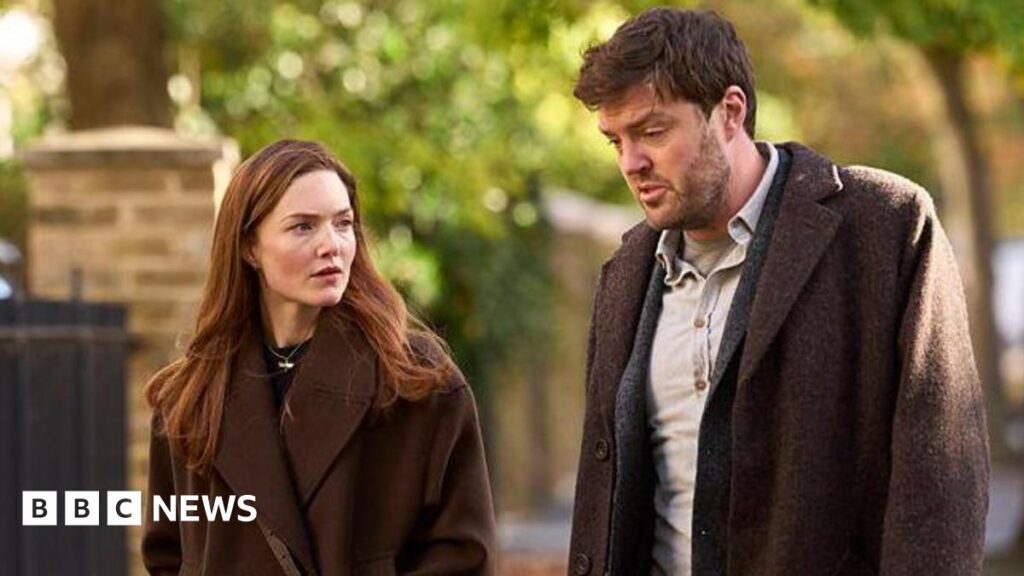 BBC films JK Rowling’s detective series Strike in Norwich Holliday Grainger acting as Robin Ellacott and Tom Burke as Cormoran Strike in BBC drama Strike. Both actors are walking outside and are wearing brown coats. They are not looking at the camera.