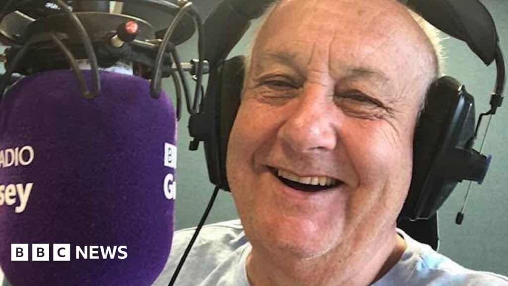 John Randall sitting behind the microphone in the BBC Guernsey studios with a big smile on his face.