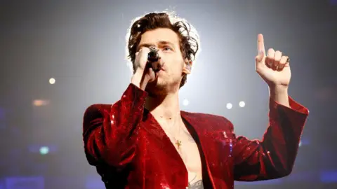 JMEnternational/Getty Images Harry Styles sings into a microphone while performing at the Brit awards in 2023. He has a red, sequinned jacket on and is pointing one finger up towards the ceiling.