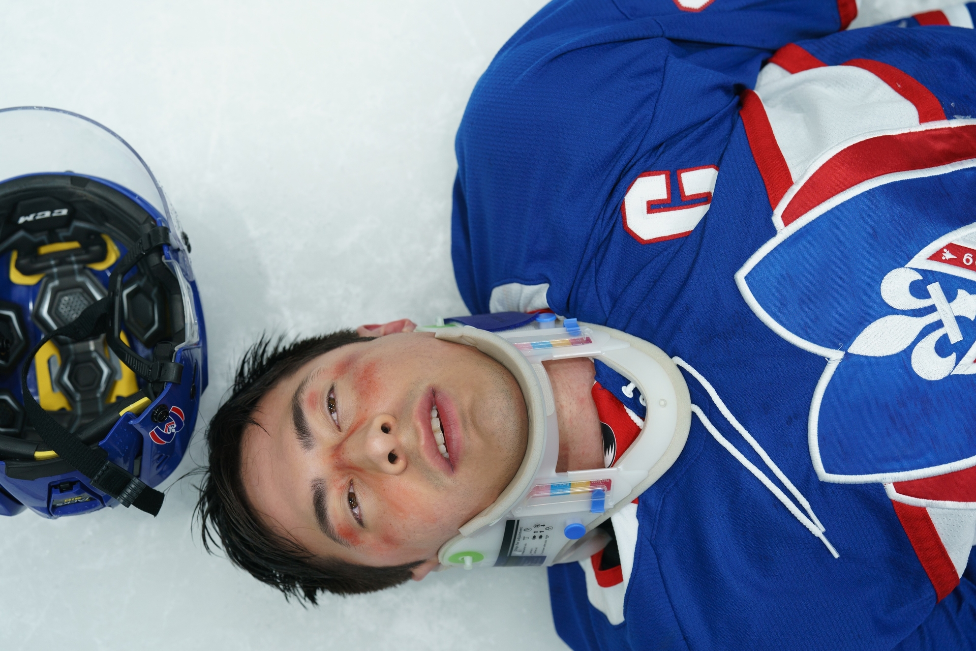 Hockey player Shane Hollander, wearing a blue jersey and neck brace, lies on the ice next to his helmet.