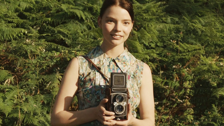 Anya Taylor-Joy's Allie stands holding a camera in front of some ferns in Marrowbone