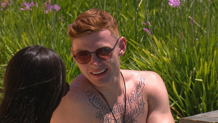 Jack Keating wearing sunglasses and topless talking to someone out of shot of the camera in the outside area of the Love Island villa.