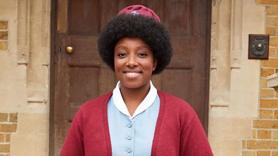 Renee Bailey in character as Nurse Joyce Highland. Wearing a midwife uniform, she stands on the steps to Nonnatus House.
