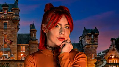 A woman in an orange top rets her head on her hand as she looks to camera, set against the backdrop of a castle by night