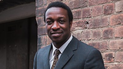 Close-up of a man wearing a suit and tie, stood outside and leaning against a brick wall.