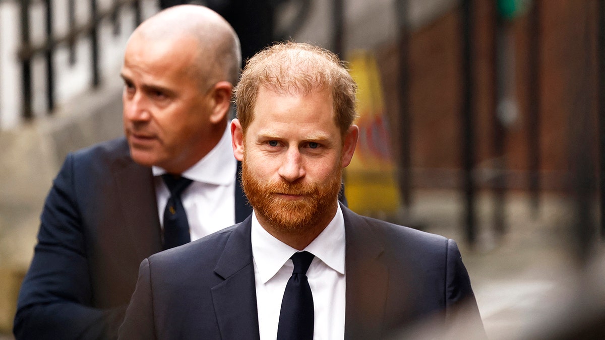 A close-up of Prince Harry walking ahead in a dark suit as his body guard keeps a watchful eye.