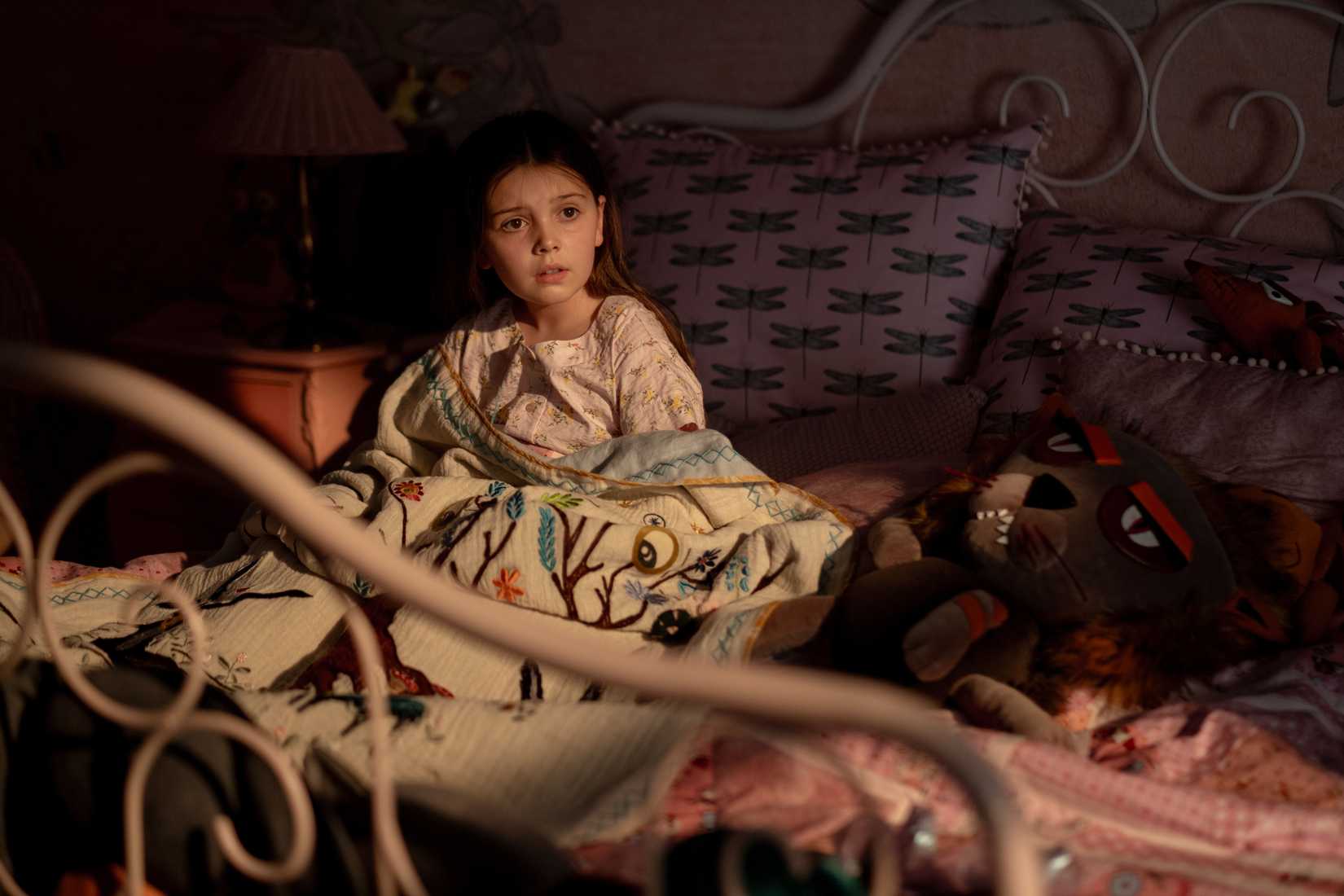 An image from Bryan Fuller's Dust Bunny featuring Sophie Sloan as Aurora. Aurora sits in her pyjamas on her bed, surrounded by her toys and bed pillows.