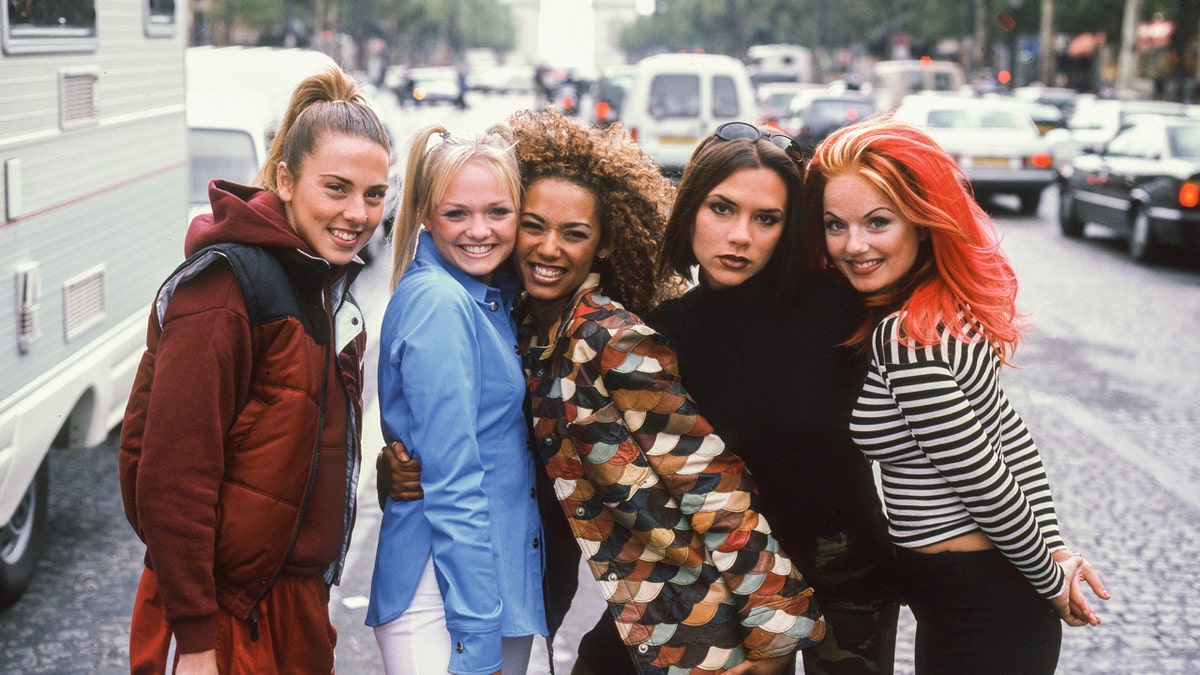 The Spice Girls posing for a portrait on a city street with the Arc de Triomphe in the background.