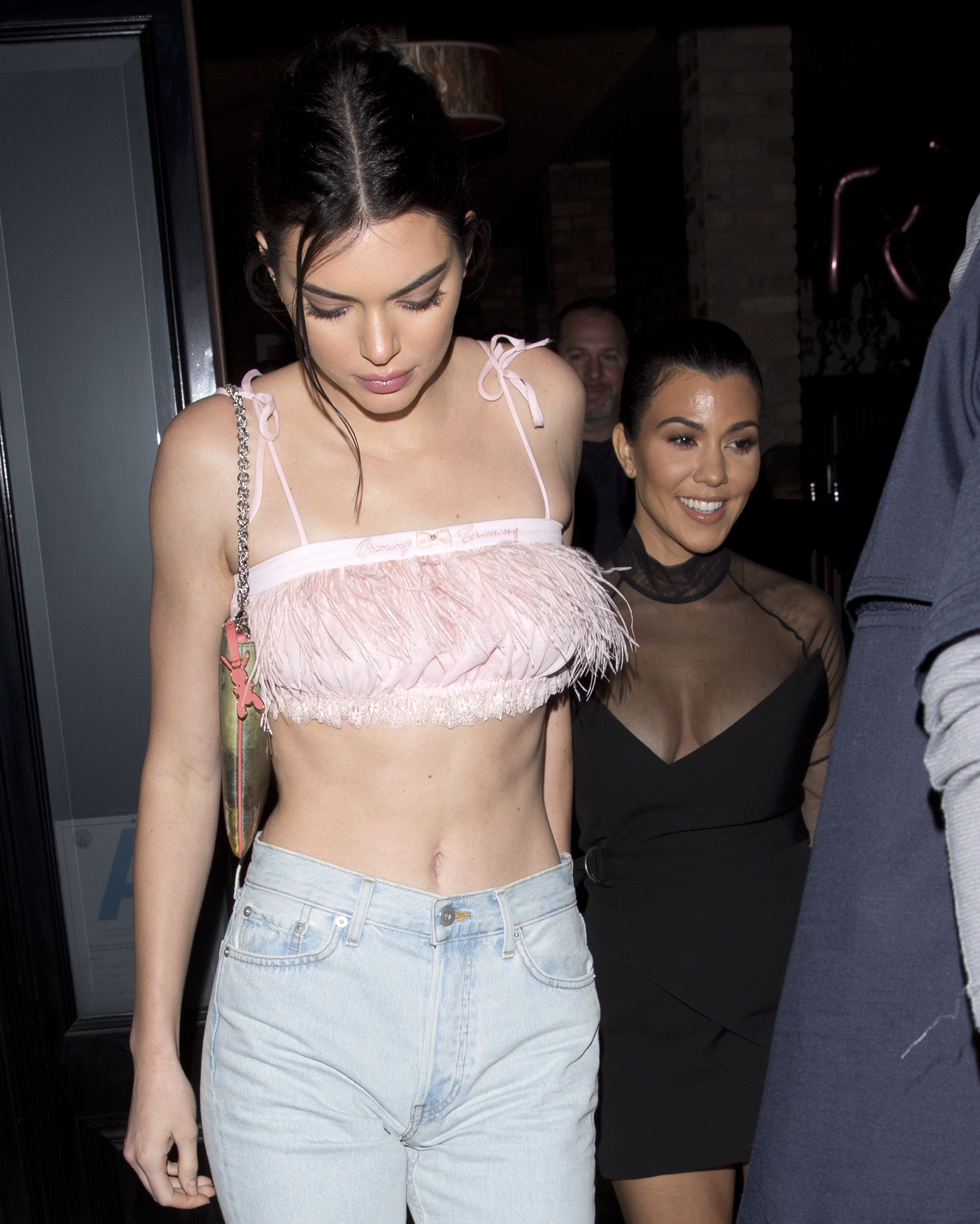 Kendall Jenner in a pink feathered crop top and light jeans with Kourtney Kardashian in a black dress, leaving a restaurant at night.