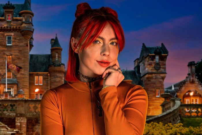 A woman smiling ahead with one hand resting on her cheek. She is wearing an orange turtleneck and has bright red hair.