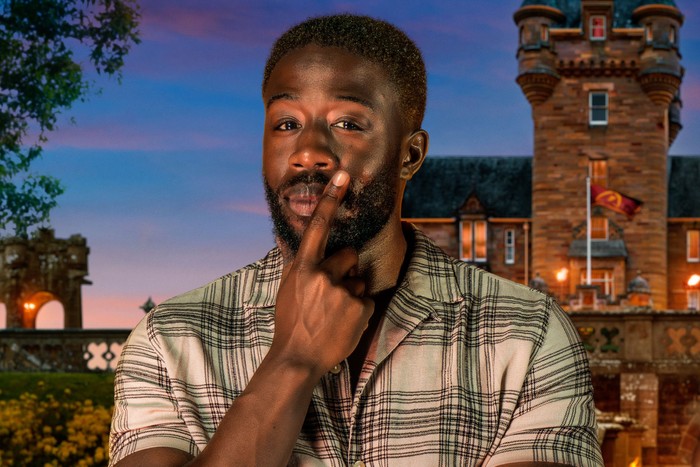 A close up of Ross, wearing a plaid patterned shirt, resting a finger on his face.