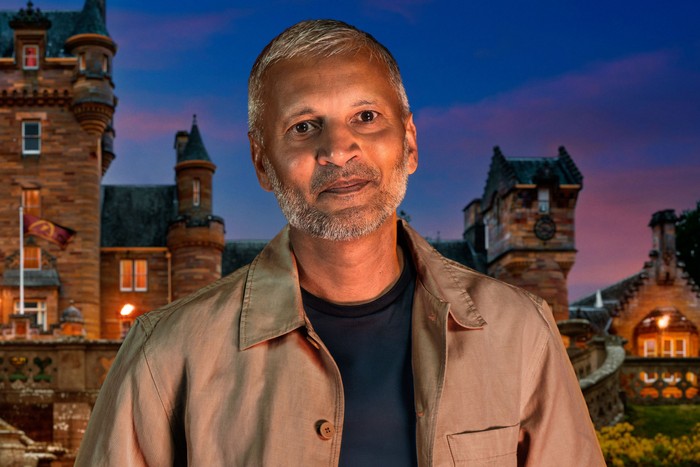 A man wearing a dark blue top and brown jacket, looking ahead with a slight smile.