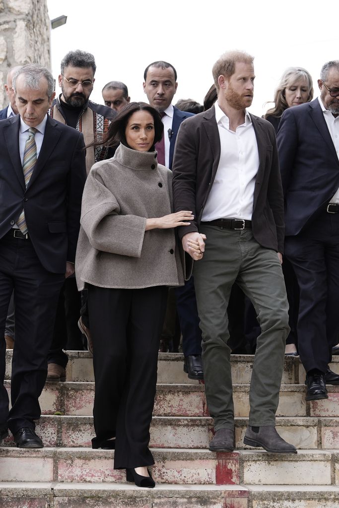 Meghan in taupe funnel-neck jacket descending steps with prince harry with people behind them