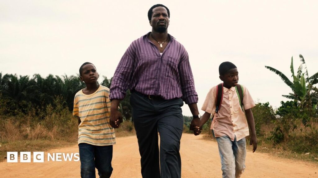 Actor Sope Dirisu walks down a dusty road whole holding the hands of young actors Godwin Chiemerie Egbo and Chibuike Marvellous Egbo. Sope is wearing a purple striped shirt and jeans. The boys are wearing jeans and T-shirts.