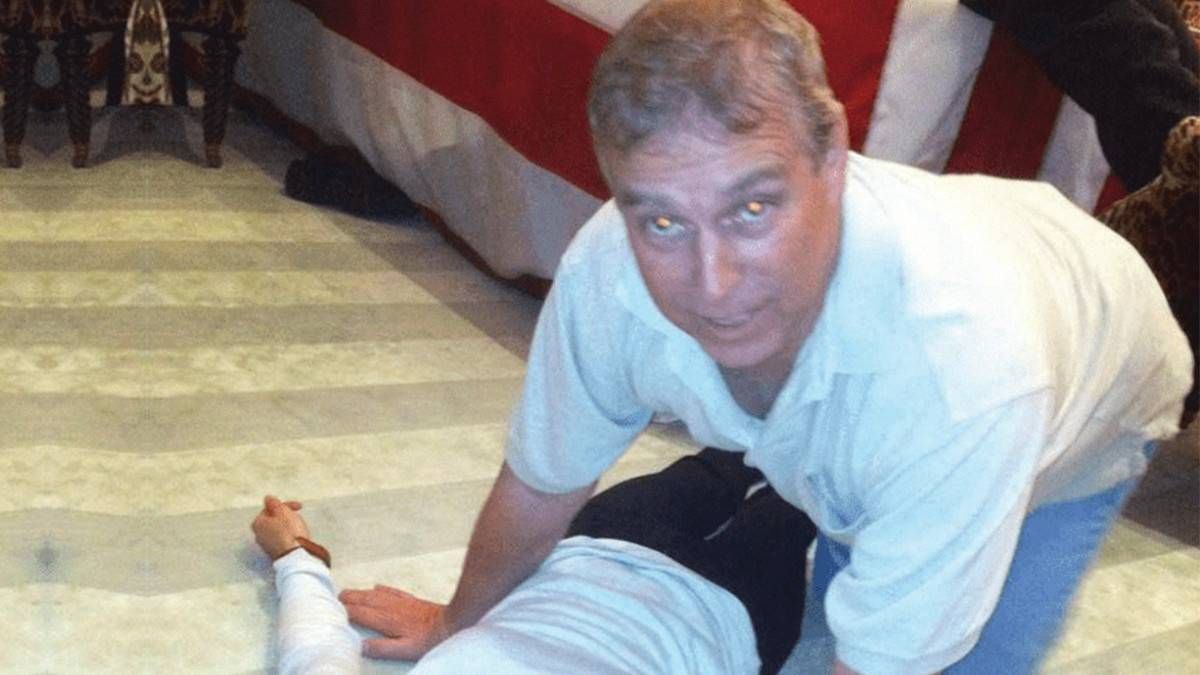An individual, dressed in a white polo shirt and black shorts, is kneeling on a tiled floor, providing assistance to another person lying on the ground. The person receiving aid appears to have an injury, as indicated by a visible bandage. The scene takes place in an indoor setting, with a bed adorned with a red and white blanket visible in the background.