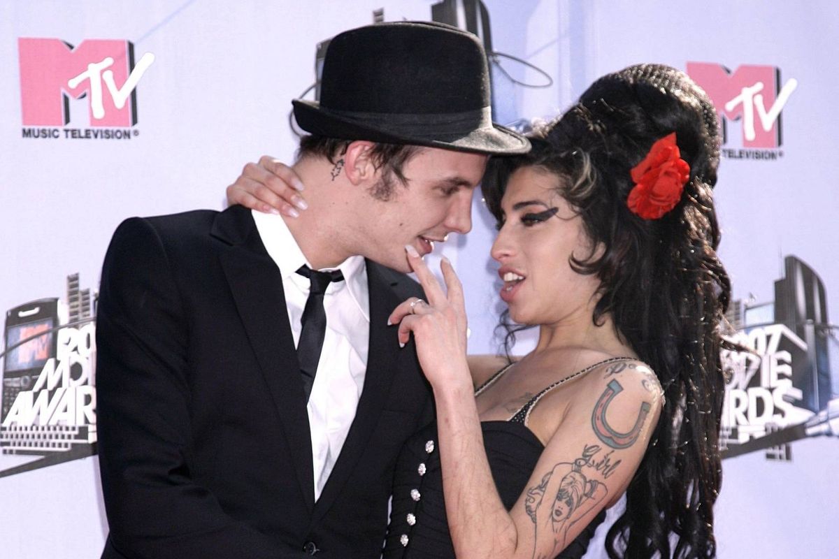An image depicts a couple standing closely together, appearing to engage in a moment of affection or conversation. The man is dressed in a black suit and hat, while the woman has long hair and is wearing a black dress. They are positioned on a stage, with a backdrop featuring logos and some graphic elements.