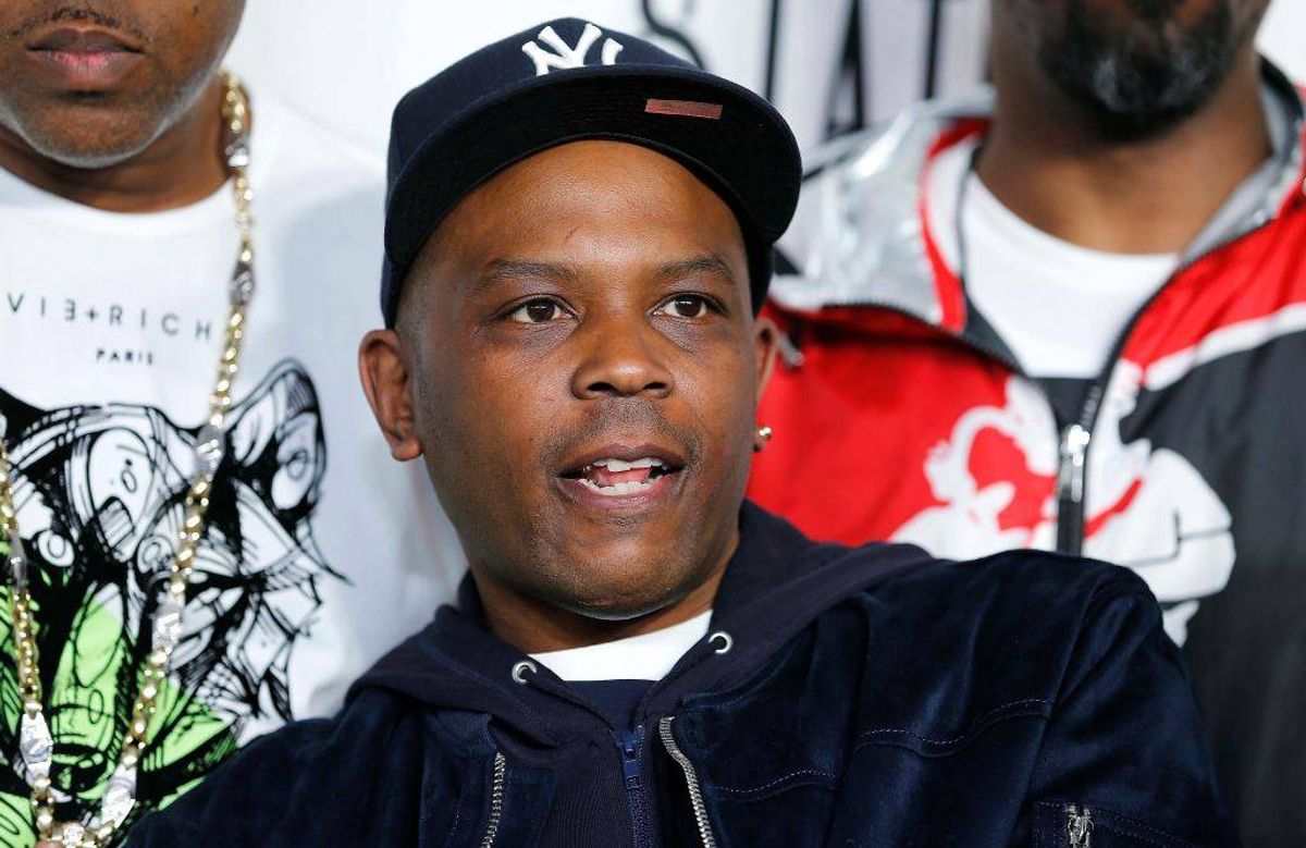 A formal photograph features an individual dressed in a black cap and jacket, standing with a slight smile, with two other individuals in the background wearing distinctive white and red apparel.