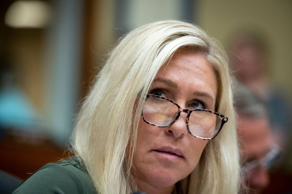 An adult woman with blonde hair and glasses is sitting at a desk in a conference room, looking down with a serious expression.