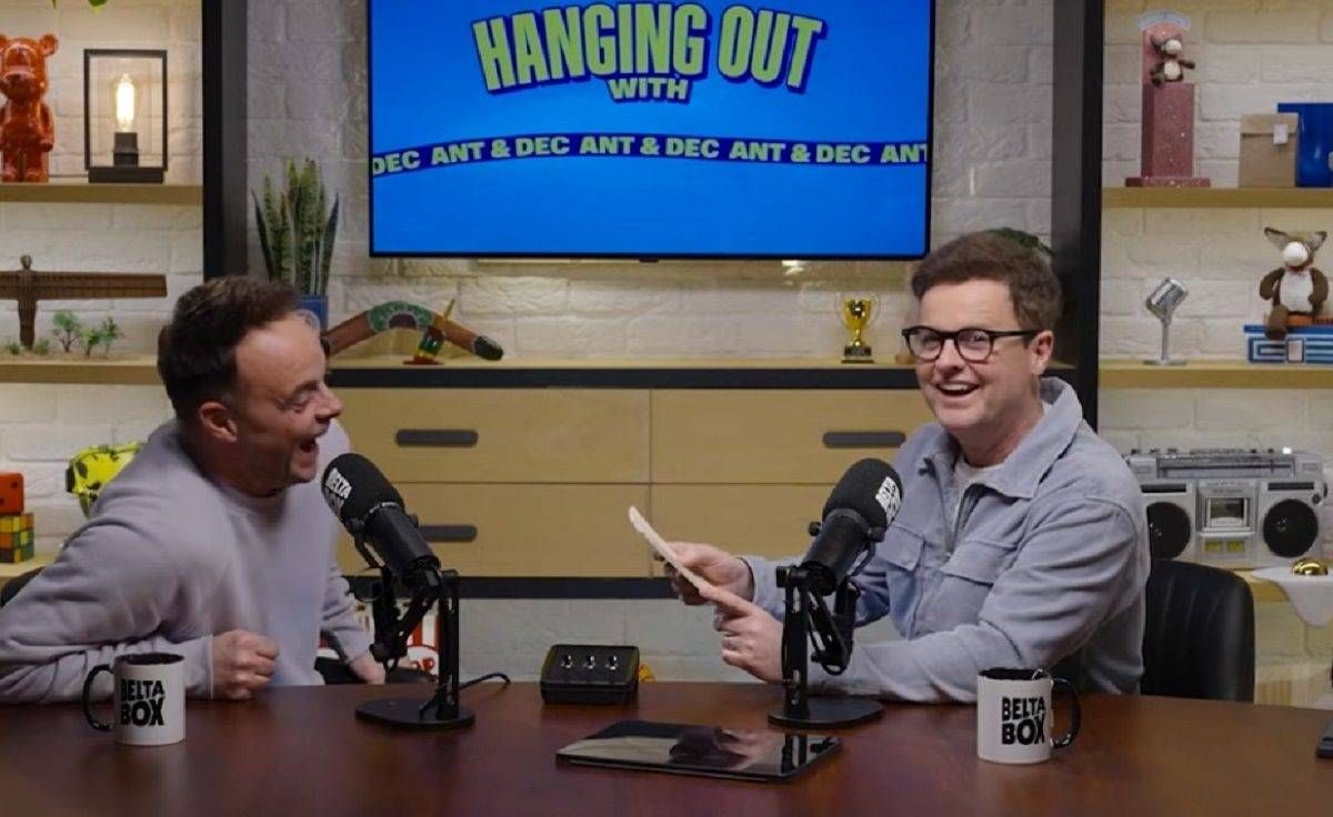 Two individuals are seated at a table, engaging in a conversation while positioned in front of microphones. The setting appears to be a studio or professional environment, with a television screen in the background displaying the title "Hanging Out." Various items such as coffee mugs, a speaker, and a potted plant are visible on the table and shelves.