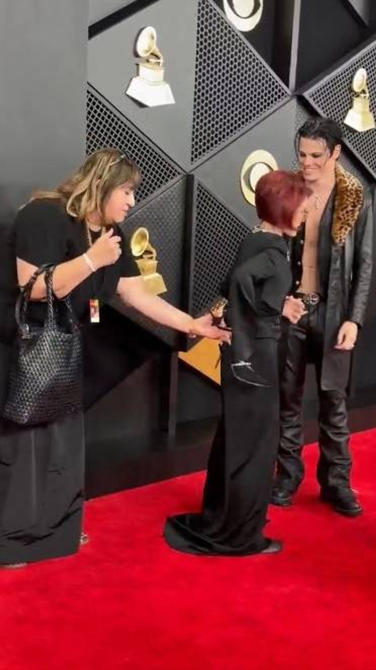 An image depicting a gathering of individuals in an award ceremony setting. One individual is holding a golden trophy, and another is carrying a handbag. The backdrop features a distinctive geometric design, likely part of the venue's interior.
