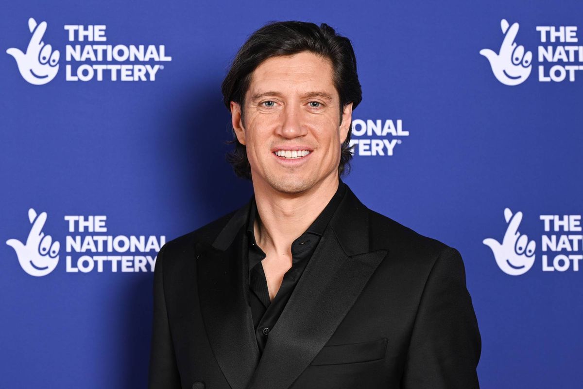 An individual dressed in a black suit stands against a backdrop featuring the word "National" prominently displayed in white, with a portion of the text obscured, suggesting a formal event setting.