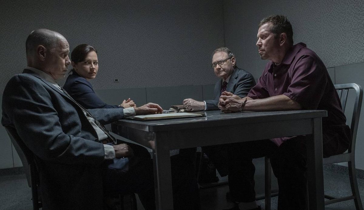 Four individuals are seated around a table in a dimly lit room, engaged in a discussion. The person on the right wears a maroon shirt, while the individual on the left is dressed in a blue suit. The central figure is wearing a dark jacket and has glasses on. A document lies on the table before them.