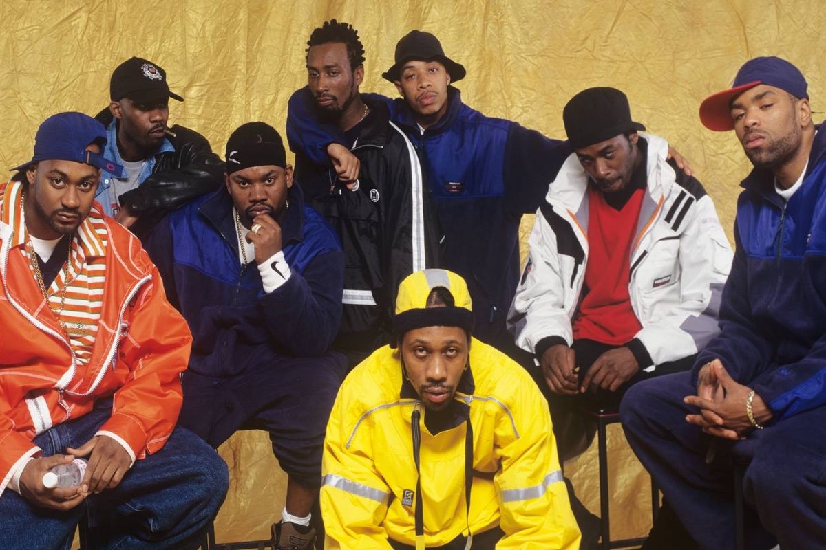 A group of individuals dressed in various jackets and hats, posing together against a yellow backdrop, with some members seated on chairs.