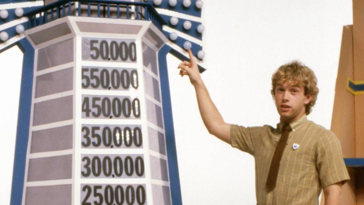 An individual, dressed in a light-colored shirt and a tie, is gesturing towards a large numerical display mounted on a building. The display features a sequence of numbers ranging from 250,000 to 550,000, possibly indicating a countdown or a financial metric.