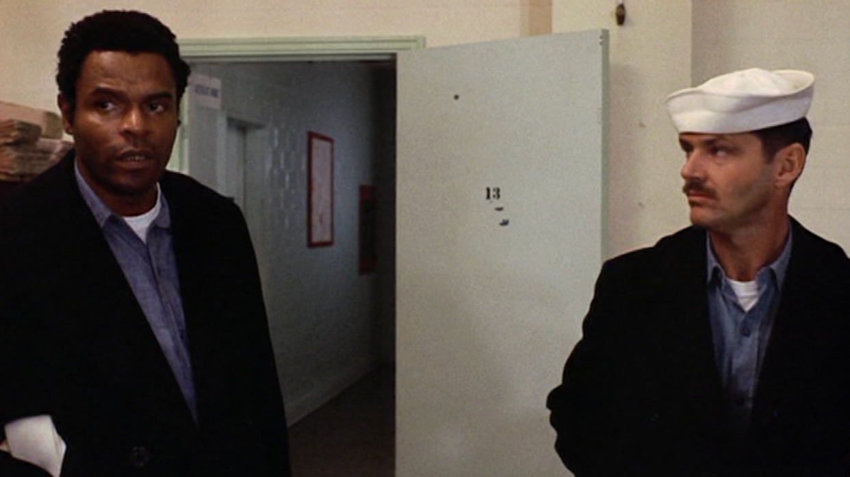 Two men stand in a hallway; one wears a dark jacket and blue shirt, the other wears a white sailor hat and uniform. An open white door with the number 13 is in the background. Both men look serious.