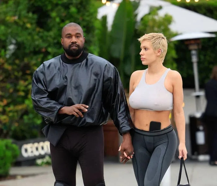Two people walking hand in hand. The man wears a loose, shiny top and fitted pants. The woman sports a cropped top and fitted pants, holding a small bag