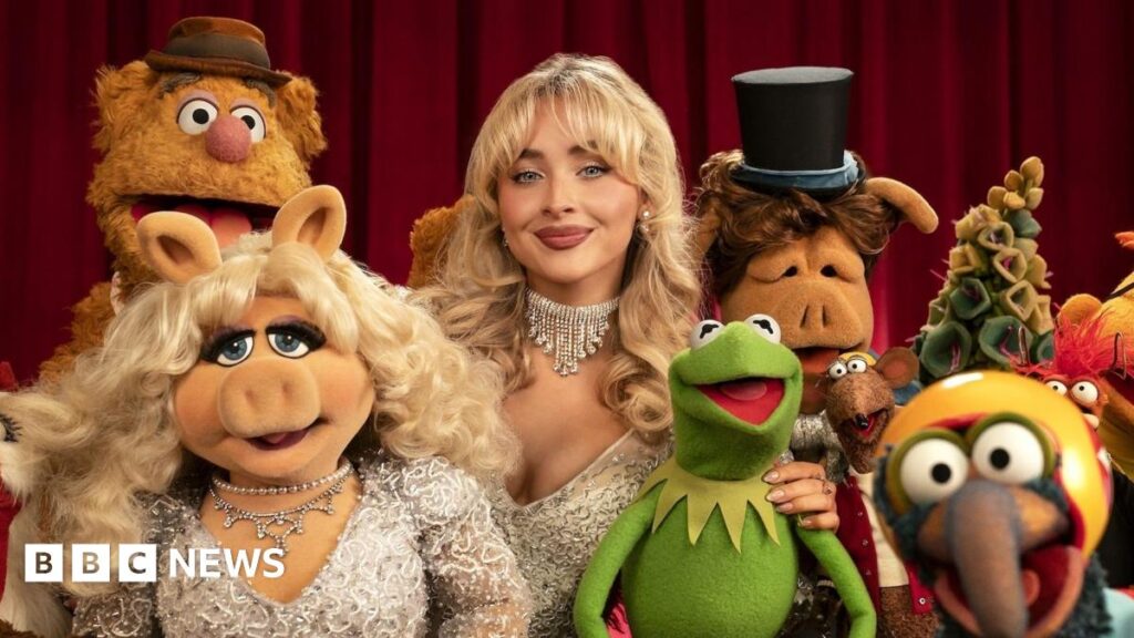 Sabrina Carpenter, centre, stands against a red velvet curtain surrounded by Muppet Show characters including Kermit the Frog, who she has her hand around, and Miss Piggy, who is wearing the same beaded ivory dress and diamond necklace as Carpenter