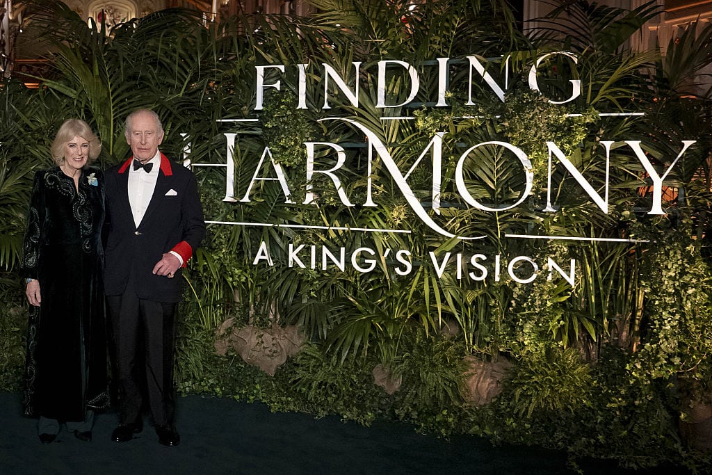 Two people stand beside a large sign that says Finding Harmony: A King's Vision, which is in front of large green plants.