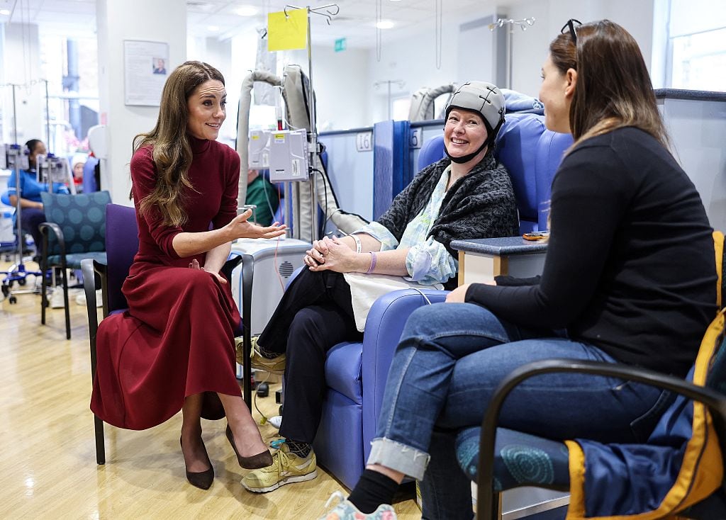 Two people talk with another person sitting in a chair receiving medical treatment.
