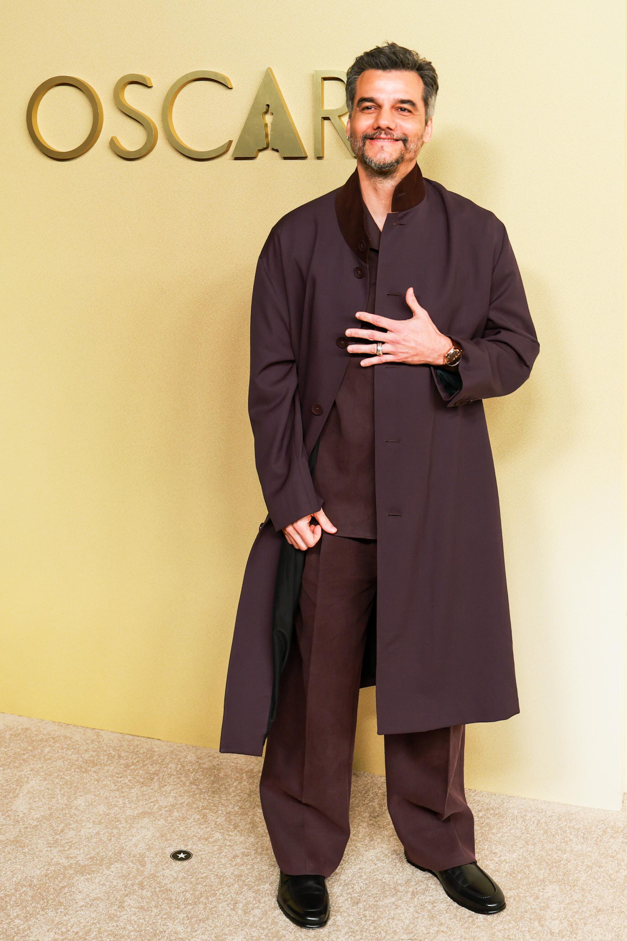 Wagner Moura, wearing a burgundy coat that matches his shirt and pants, poses on the beige carpet. 