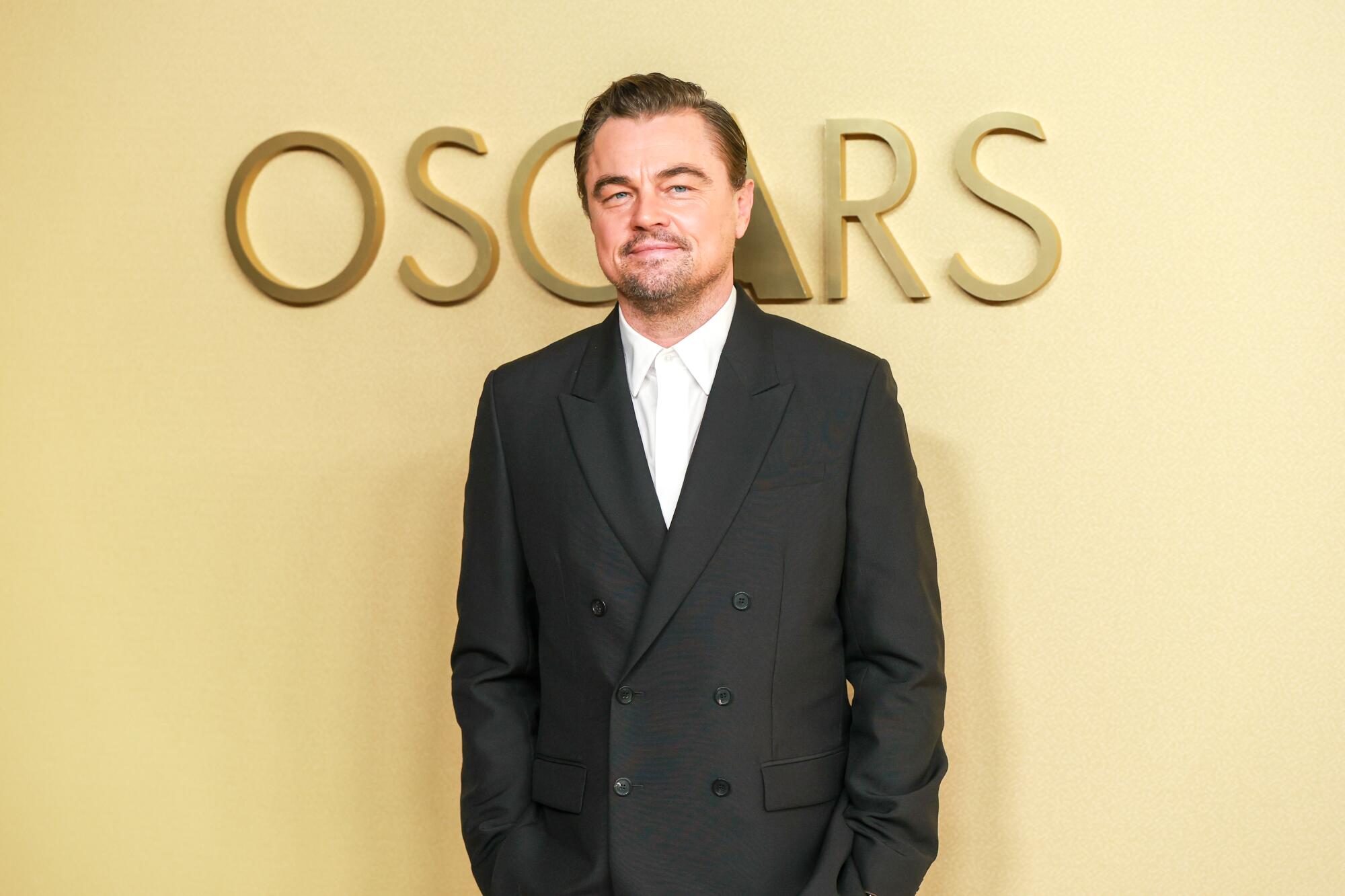 Leonardo DiCaprio, wearing a black suit and white tie, poses on the beige carpet. 