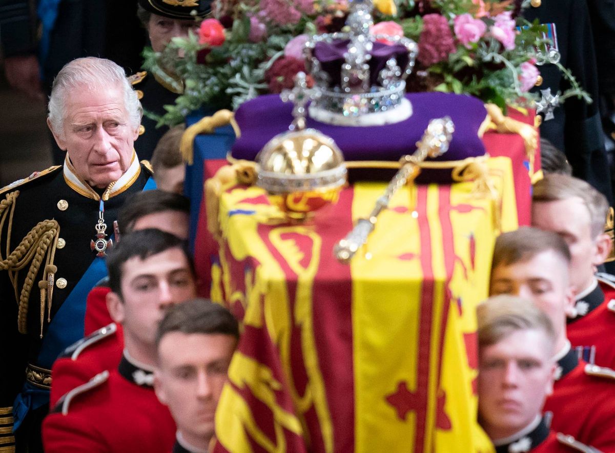 A solemn procession with a coffin draped in a yellow and red cloth, adorned with a crown and surrounded by individuals in military uniforms.
