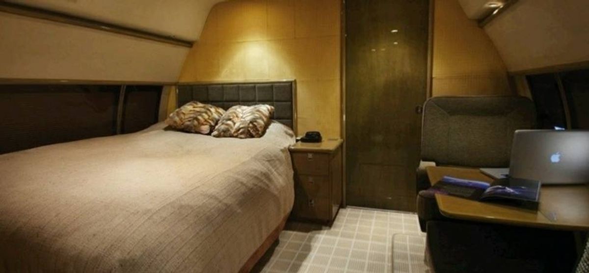 The interior of a bedroom showcases a bed adorned with two decorative pillows and a white bedspread. Adjacent to the bed, there is a nightstand housing a lamp. The room features yellow walls and a white carpeted floor. A desk with books and a chair is positioned in the right corner of the room.