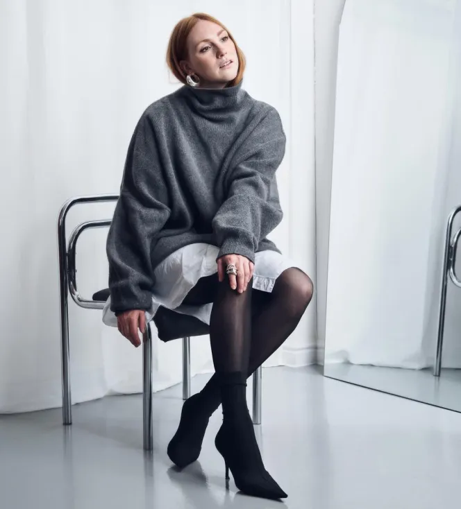 Aimee Croysdill sitting on a chrome chair, wearing a gray turtleneck sweater over a white dress, black tights, and black high heels, looking up and to the right.