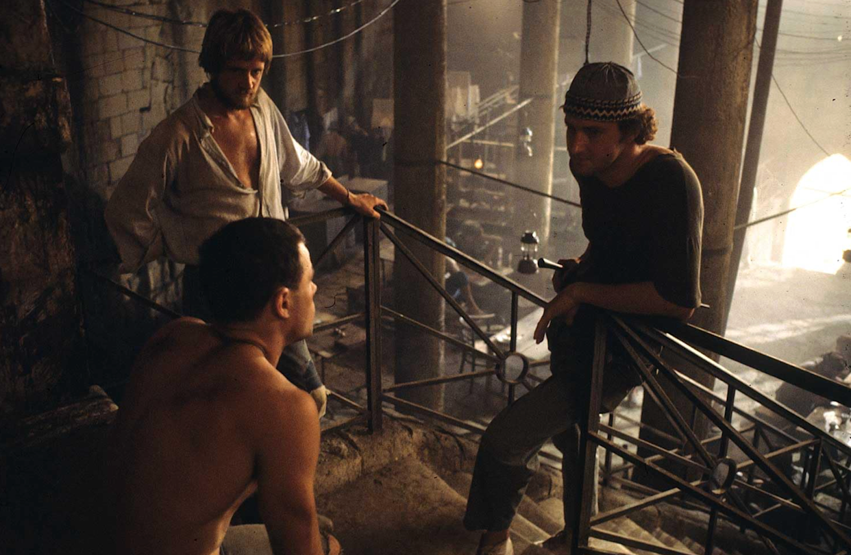 Three men are talking on a dimly lit, industrial staircase. One is shirtless, another wears a white shirt, and the third wears a dark shirt and patterned hat. The background is filled with columns, wires, and a hazy atmosphere.