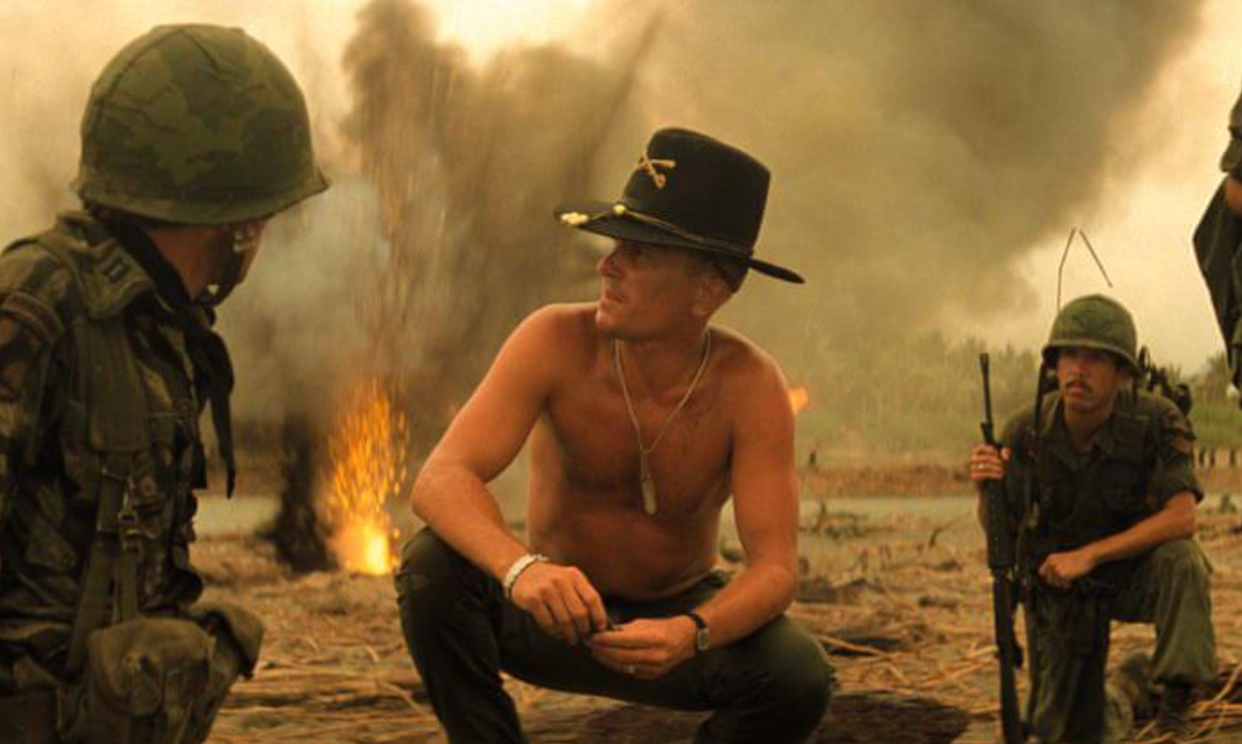 Three soldiers in military gear kneel and crouch on a smoky battlefield; one is shirtless and wears a black cavalry hat and sunglasses. An explosion and heavy smoke rise in the background.
