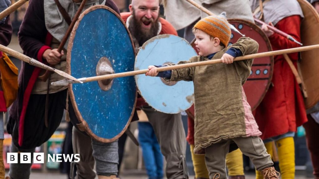 A little boy in costume wields a spear while men behind him hold large wooden shields