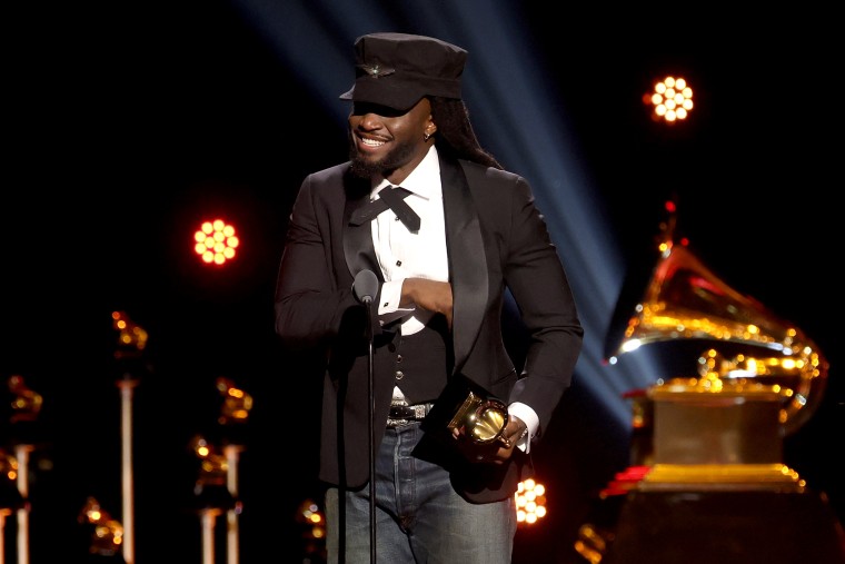 Shaboozey, wearing a black cap, tuxedo jacket and blue jeans, stands at a microphone while holding a Grammy award.