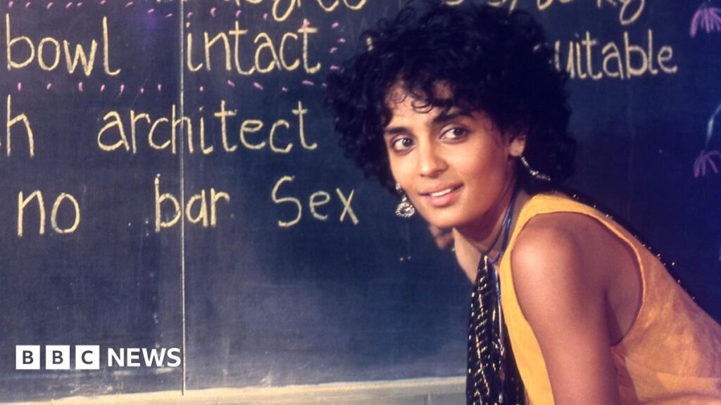 A young Arundhati Roy wearing a yellow tunic and a black scarf bends slightly in front of a blackboard in a scene from In Which Annie Gives It Those Ones