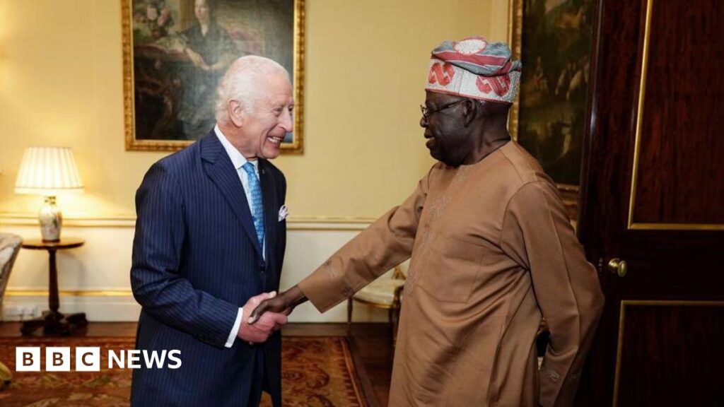 King Charles, dressed in a navy pin stripe suit, white shirt and sky blue tie, shakes hands with President Tinubu, who wears a light brown outfit and colourful kufi. They stand in a brightly lit room, with pale yellow walls adorned with gold-framed paintings and wooden floors decorated with a dark red rug.